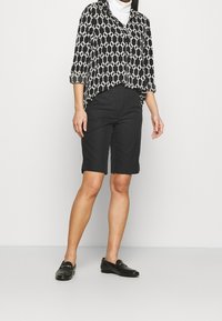 Koszula w czarno-biały wzór z podwiniętymi rękawami, w połączeniu z czarnymi szortami i czarnymi mokasynami. Gładka tkanina o luźnym kroju.