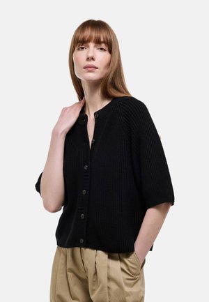Femme aux cheveux lisses châtain clair, portant un cardigan en maille noir boutonné et un pantalon plissé beige, une main dans la poche et l'autre sur l'épaule.