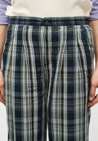 Άτομο που φοράει παντελόνι με καρό μοτίβο σε σκούρο μπλε, άσπρο και κίτρινο, με κουμπωτή μέση και τα χέρια ακουμπισμένα στις πλευρές τους.