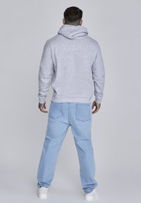 Felpa grigia da uomo con cappuccio e cordoncino, abbinata a jeans azzurri a gamba larga. L'outfit presenta una texture liscia e un design rilassato.