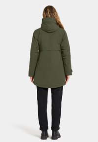 Vrouw met lang haar, gekleed in een olijfgroene gewatteerde jas met capuchon, zwarte broek en zwarte laarzen, staand met de rug naar de camera op een effen achtergrond.