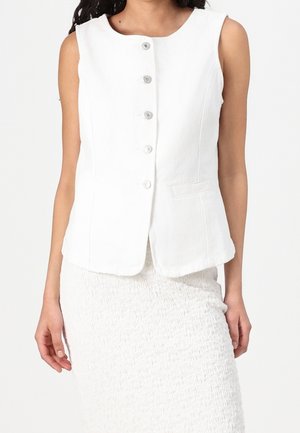Femme portant un gilet blanc sans manches boutonné associé à une jupe blanche texturée sur un fond uni.