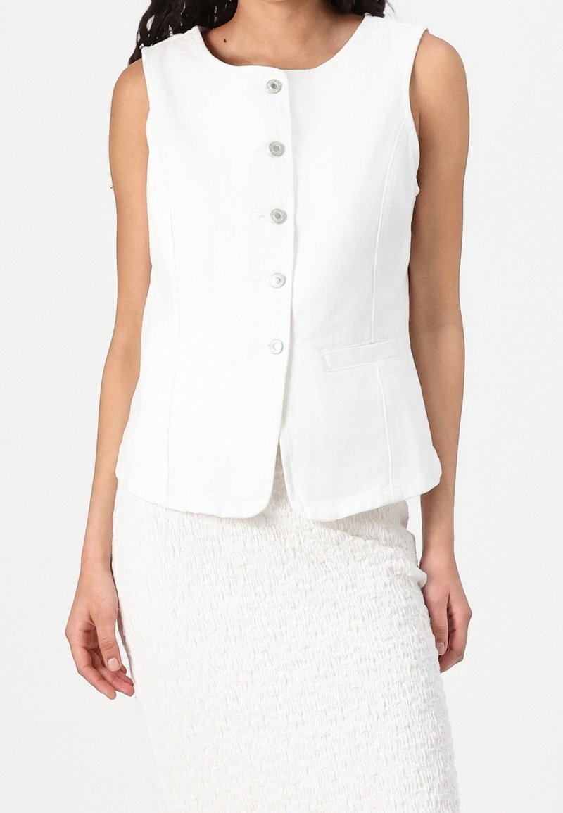 Femme portant un gilet blanc sans manches boutonné associé à une jupe blanche texturée sur un fond uni.