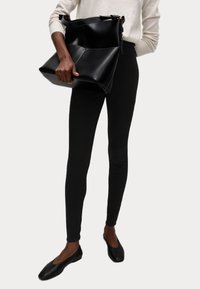 Bolso de cuero negro con un diseño estructurado, que presenta un asa superior y costuras visibles. Combinado con leggins negros y zapatos planos.