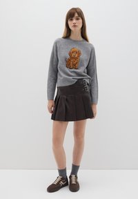 Grauer Pullover mit braunem Welpenmotiv, kombiniert mit einem plissierten braunen Lederrock mit seitlichen Schnallen. Braune Sneakers und graue Socken runden den Look ab.