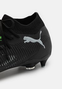 Botín de fútbol negro con una parte superior texturizada, que presenta un cuello de tobillo de punto, acentos verdes y un prominente logo plateado de Puma cerca del talón.