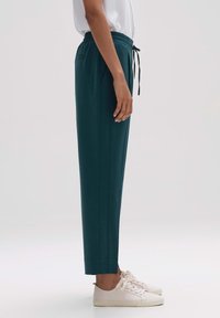 Pantalones jogger en color verde azulado con cintura elástica y cordón, con bolsillos laterales y un corte ajustado, combinados con zapatillas de color claro.