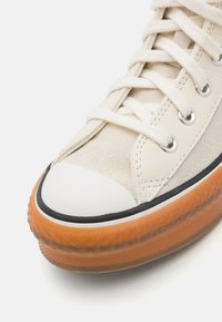 Baskets en suède et cuir beige avec un embout en caoutchouc blanc, des accents noirs et une semelle translucide orange. Elles présentent des œillets ronds et des lacets plats.