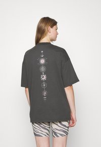 Tmavě šedé oversized tričko s grafickým potiskem geometrických a hvězdných vzorů v bílé a růžové na zadní straně. Krátké rukávy.