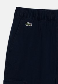 Mørkeblå bukser med elastisk linning, med sidelommer og en grønn brodert krokodille-logo på forsiden.