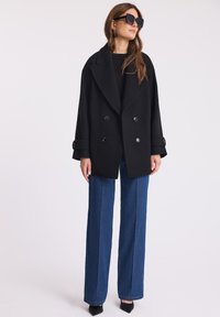 Czarny podwójnie zapięty płaszcz z dużymi klapami, z teksturowanej wełny, w połączeniu z wysokim wcięciem w niebieskich dżinsowych spódnicach i spiczastymi czarnymi szpilkami.