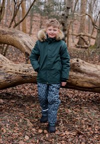 Green insulated jacket with faux fur hood, gray patterned pants, and black boots. Set in a forest with fallen leaves and a log.