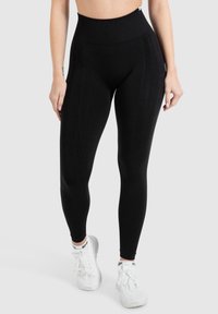 High-waisted zwarte legging van rekbaar materiaal, met een zachte textuur en subtiele zijnaaddetails, gecombineerd met witte sneakers.