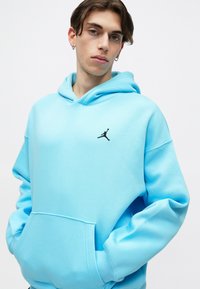 Tânăr îmbrăcat cu un hanorac albastru deschis cu un logo Jumpman negru, stând cu mâinile în buzunare pe un fundal alb simplu.