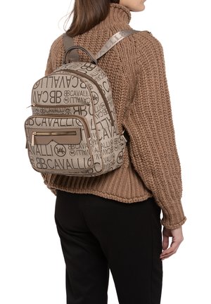 Beige rugzak met logo-print en een gladde textuur, met een ritsvak aan de voorkant en verstelbare schouderbanden, gedragen op een schouder gekleed in een bruine trui.