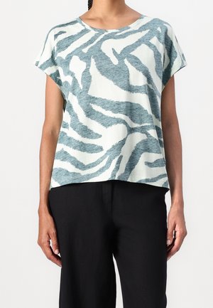 Personne portant un haut ample à manches courtes avec des rayures zèbre abstraites bleues et blanches et un pantalon noir sur un fond uni.