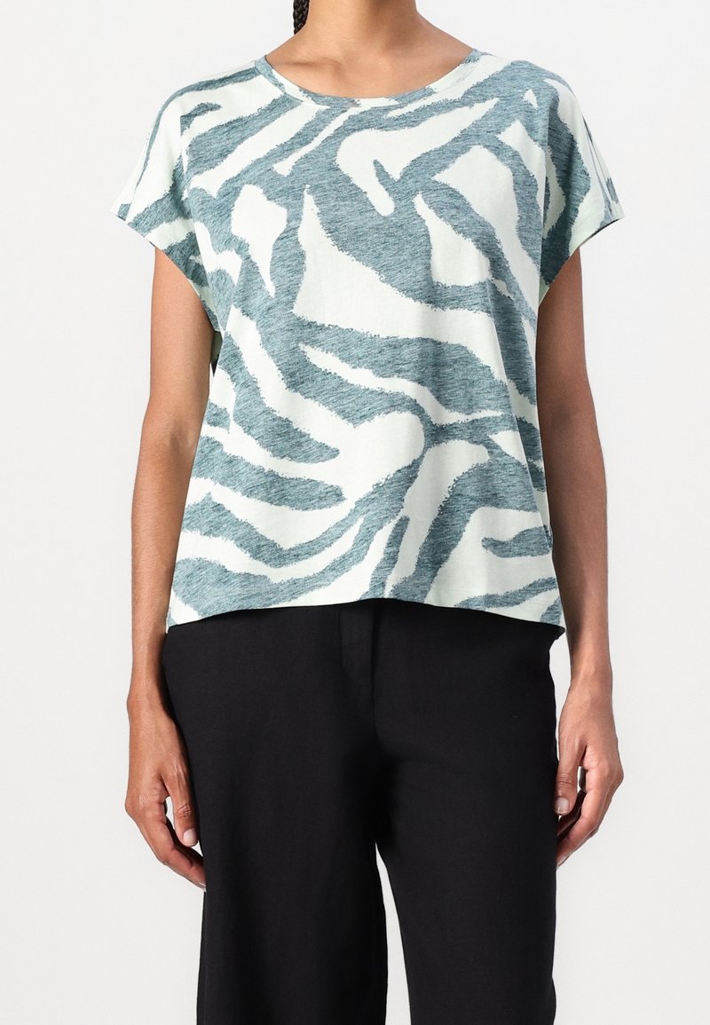 Personne portant un haut ample à manches courtes avec des rayures zèbre abstraites bleues et blanches et un pantalon noir sur un fond uni.