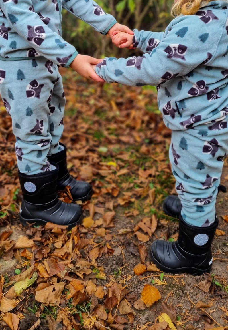 Blå fleecepyjamas med tvättbjörnstryck; svarta vattentäta stövlar; två barn som håller handen bland fallna höstlöv.