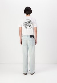 T-shirt blanche avec un logo circulaire noir "Santa Cruz" au dos, associée à un jean bleu clair à coupe ample et des chaussures foncées.