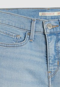 Jean en denim bleu clair en gros plan montrant la poche avant, la boucle de ceinture, le bouton en métal et l’étiquette indiquant "318 Shaping Wide Leg 27".