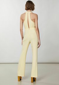 Mono sin mangas amarillo con un escote en forma de ojal en la parte posterior, cintura ajustada, piernas acampanadas y un detalle de cremallera. Tejido texturizado con un brillo sutil.