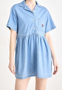 Ljusblå denimklänning med kragardesign, korta ärmar, knapplist fram och samlad midja. Har en bröstficka med varumärkeslogotyp.