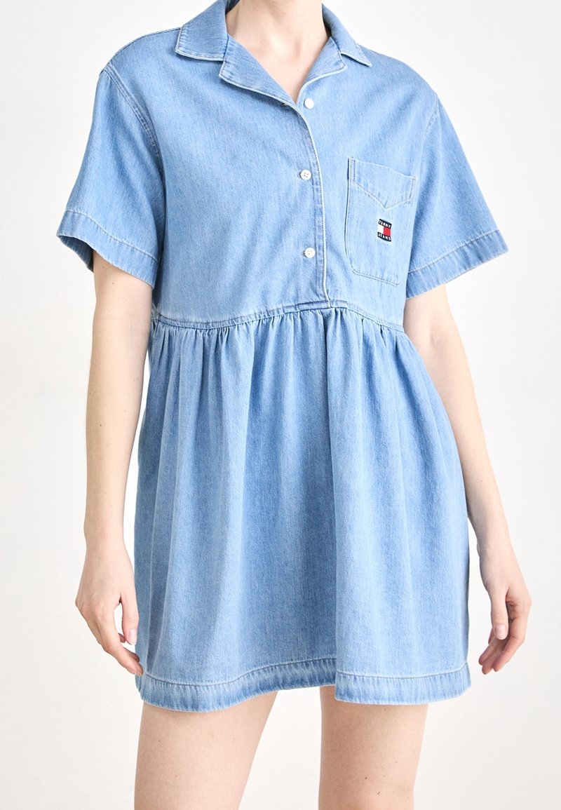 Ljusblå denimklänning med kragardesign, korta ärmar, knapplist fram och samlad midja. Har en bröstficka med varumärkeslogotyp.