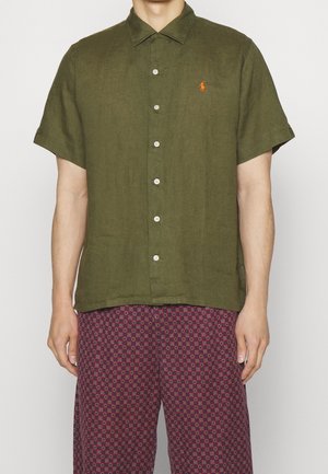 Homme portant une chemise boutonnée à manches courtes de couleur vert olive avec un logo brodé orange et un pantalon à motifs rouges et noirs.