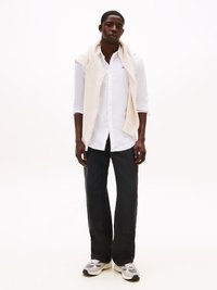 Camicia bianca a maniche lunghe, dolcevita color crema appoggiata sulle spalle, pantaloni neri a gamba larga, sneaker grigie con dettagli neri, sfondo neutro.