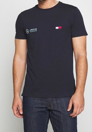 Man draagt een zwart AMG Petronas Motorsport T-shirt en donkerblauwe spijkerbroek, staand met ontspannen handen langs het lichaam.