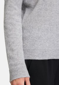 Szürke gyapjúpulóver bordázott mandzsettákkal és szegéllyel, puha textúrával és laza szabással, részben fekete nadrág háttér előtt látható.