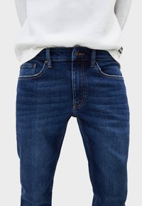 Mörkblå denimjeans med rak passform, femficksdesign och synliga sömmar. Har en metalldetalj som knäppning och ett stadigt midjeband.