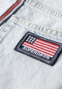 Le macaron Superdry sur le denim bleu clair présente un drapeau américain brodé avec des rayures rouges et blanches, entouré d'une couture noire.
