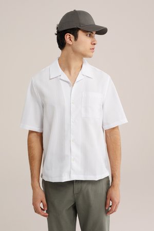 Junger Mann, der eine graue Kappe, ein weißes kurzärmliges Hemd mit Knöpfen und olivgrüne Hose trägt und nach rechts blickt, vor einem schlichten Hintergrund.