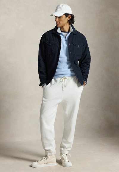 Moški z belo kapo, črno jakno, svetlo modrim puloverjem s četrtinskim zadrgo, belimi trenirkami z vrvico in bež visokimi supergami, stoji z rokami v žepih.