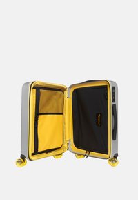 Valigia rigida grigia con interno giallo. Presenta una tasca in rete, rivestimento nero e quattro ruote gialle per la mobilità.