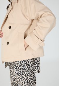 Beige trench coat with a double-breasted design, featuring black buttons, side pockets, and adjustable cuff straps. Pairs with leopard print pants.