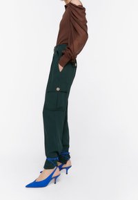 Hnědý dlouhý rukáv s nařasenými detaily na ramenou, spárovaný se zelenými cargo kalhotami s knopovými kapsami. Modré lodičky s špičatou špičkou dokončují vzhled.