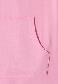 Różowy materiał z prostokątną przednią kieszenią z estetycznym przeszyciem i zaokrąglonym otworem po prawej stronie.