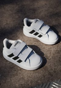 Bijele sportske tenisice sa sivim detaljima, s tri čičak-trake i crnim bočnim prugama. Glatki materijal i zaobljeni vrh.