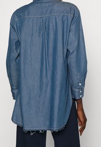 Denim overhemd in middenblauw, met lange mouwen, knoopmanchetten en een licht afgeronde zoom. De stof lijkt zacht met een lichte structuur.