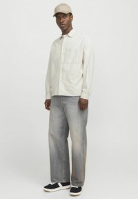 Helles graues Button-up-Hemd mit einer Brusttasche, kombiniert mit locker sitzenden hellblauen Jeans und schwarzen Sneakern mit einer weißen Sohle. Beiger Hut.
