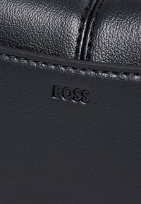Borsa nera in pelle con superficie texturizzata, che presenta il logo "BOSS" impresso e dettagli di cucitura discreti. Design compatto, materiale resistente.
