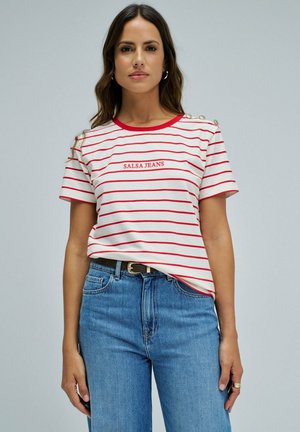 Mujer con camiseta de rayas blancas y rojas "Salsa Jeans" y vaqueros azules, de pie frente a un fondo claro y liso.