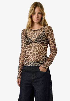 Mujer con un top transparente de manga larga con estampado de leopardo sobre un sujetador negro y jeans de mezclilla oscuros de talle alto, con una mano en el bolsillo.