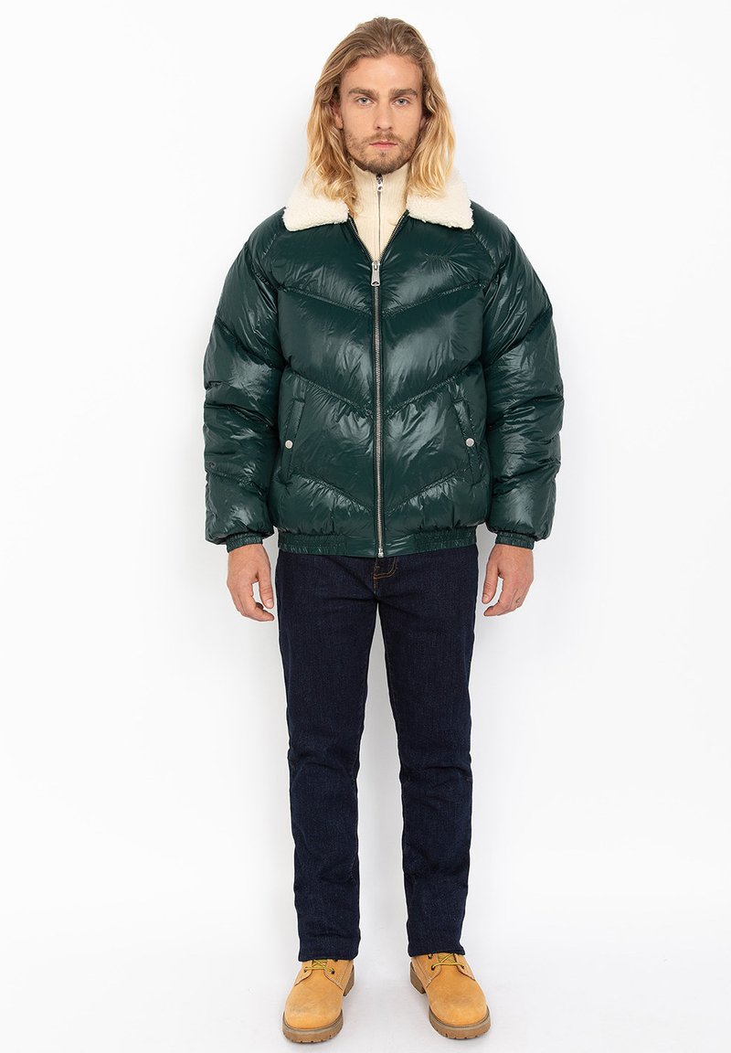 Green puffer jacket with a shiny finish, cream faux fur collar, zip closure, and elastic cuffs, paired with dark jeans and tan boots.