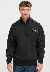 Sudadera negra con cuello de media cremallera, que presenta las letras "A.R.C." en blanco en el lado izquierdo del pecho. Confeccionada con una suave tela texturizada.