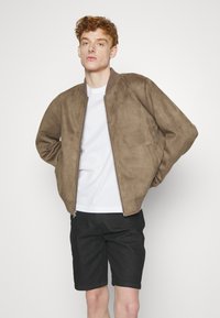 Chaqueta bomber de ante marrón con puños acanalados, cierre de cremallera y bolsillos laterales, llevada sobre una camiseta blanca y pantalones cortos negros. Fondo liso.