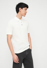 Jeune homme portant un polo en maille à manches courtes blanc et un jean noir, debout avec une main dans la poche, de profil.