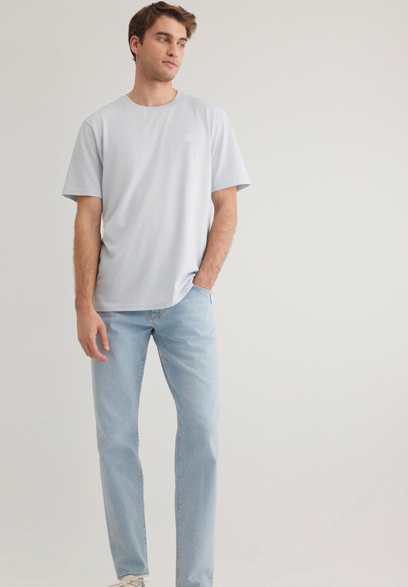 Hellblaues T-Shirt mit kurzen Ärmeln, rundem Ausschnitt und kleinem Logodetail auf der linken Brust, kombiniert mit hellblauen Jeans und weißen Sneakern.